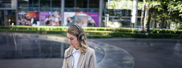 Young businesswoman sitting in city centre with laptop and headphones, joining online work meeting,...