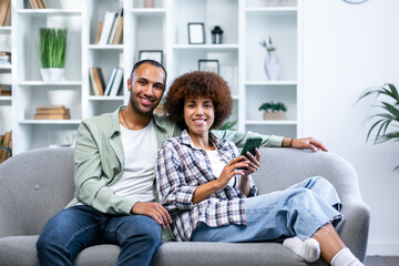 Smiling husband and wife sit on couch using smartphone, happy young couple relax on sofa look at...