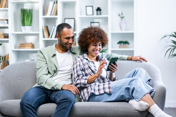 Overjoyed young couple sit rest on couch at home have fun watching video on smartphone together. Happy man and woman enjoy weekend in cozy living room - obrazy, fototapety, plakaty