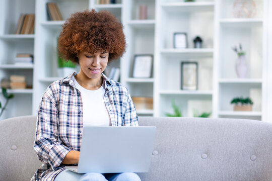 Beautiful cheerful african american curly girl using silver laptop while sitting on sofa in living room at home. - Powered by Adobe