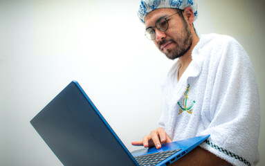 Man wearing towel and bathing cap looking blue laptop 