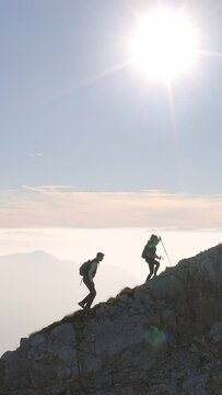 Vertical video of two silhouettes, male and female, climbing on a steep rocky mountain ridge with a blue sky and sunshine in the background, aerial side view. Challenge and goal concepts.