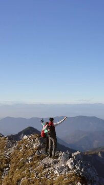 Vertical video of rear view of hugged hiker couple celebrating reaching the ridge looking at a stunning mountain range horizon, aerial view. Life, goal, success, and achievement concepts.
