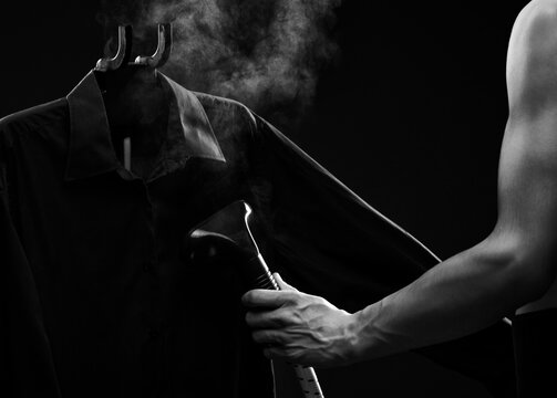 A man irons a dark shirt with a steamer on a black background.