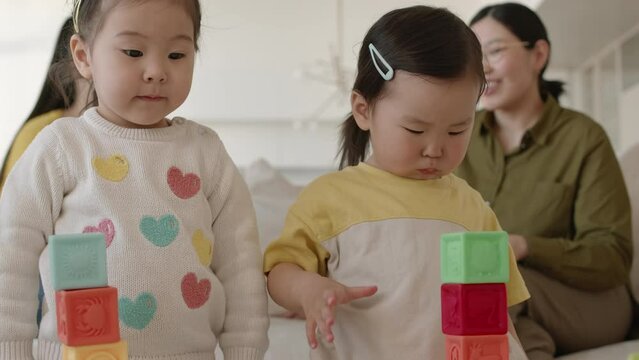 Waist up of two cute little Asian girls playing with colorful building blocks and their moms chatting on sofa in blurred background, staying in modern minimalist apartment together