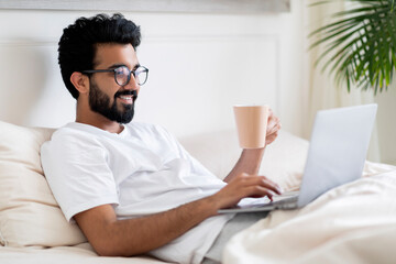 Obraz premium Young Indian Man Using Laptop And Drinking Coffee While Sitting In Bed