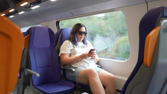 Remote view of female passenger in sunglasses using smartphone, typing chatting with friends traveling in train by window. Plus-size woman reading online news on mobile phone during train ride