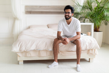 Fototapeta na wymiar Portrait Of Happy Young Indian Man Sitting On Bed In Bedroom