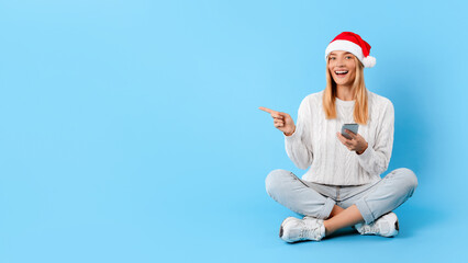 Happy caucasian lady in Santa hat using phone and pointing finger on empty space, blue background, panorama, banner