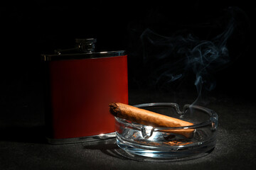 New hip flask and ash tray with cigar on dark background
