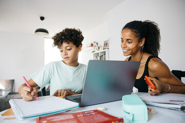 Cheerful woman helping her smiling daughter doing homework. Happy mother assisting her daughter with school homework in living room. - obrazy, fototapety, plakaty