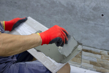 A male builder applies adhesive tile to lay it on the enclosing wall of the building from the outside. A construction contractor lays facing tiles on the fence of a private house.