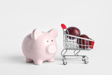 Piggy bank and shopping cart with plums on white background