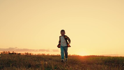 Naklejka na ściany i meble Joyful little boy running at sunset. Kid is running across field. Child boy runs through green grass in sun. Childhood dream happiness concept. Happy child playing in nature. Happy family. Dream kid
