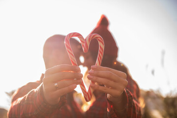 Red white candy cane is held in hands of loving couple near green market of Christmas trees