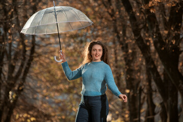 Dancing pretty girl with long hair walks through autumn park. Young woman in blue sweater with transparent umbrella