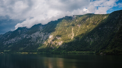 A mountain on the shore of the alpine lake Hallstätter See illuminated by the afternoon sun shining through the rain clouds. A house on the edge of the lake.