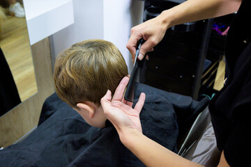 A little cute boy sits in a hairdresser's at the stylist's, a schoolchild is getting hair cut in a beauty salon, a child at a barbershop's, a short men's haircut - obrazy, fototapety, plakaty