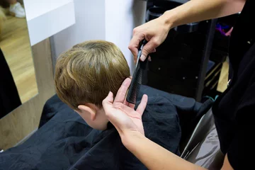 Foto auf Glas A little cute boy sits in a hairdresser's at the stylist's, a schoolchild is getting hair cut in a beauty salon, a child at a barbershop's, a short men's haircut © Miri García