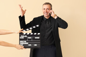 Mature actor talking by mobile phone and hands with clapperboard on beige background