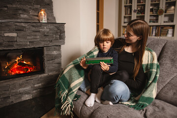 Obraz premium Happy mother and child are playing with a toy train near the fireplace. Mother and child sitting and playing on sofa near fireplace. Single parenting. Mom and baby. Parent and little kid relaxing.