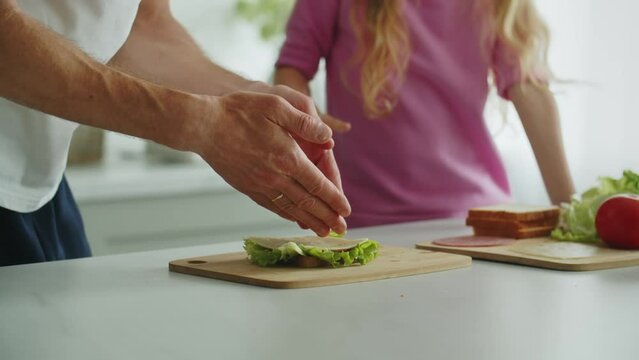 Little smiling daughter stands in the kitchen at the table and helps her father to prepare and fold a sandwich for breakfast, lunch or work in playful mood. Demonstration of careness and healthy