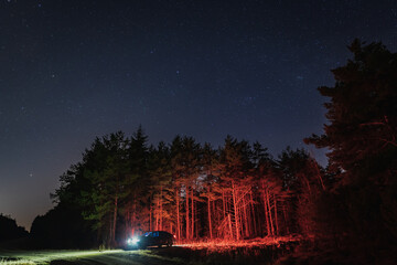 A car with headlights on in the forest at night with a starry sky.