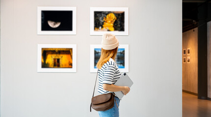 Young person at photo frame hold digital book leaning against at show exhibit artwork gallery, Asian woman holding tablet at art gallery collection in front framed paintings looking pictures on wall - obrazy, fototapety, plakaty