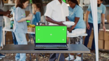 Digital laptop with blank green screen display is placed on table while volunteers help the less fortunate. Wireless computer in the foreground with isolated chromakey mockup template. Tripod shot. - obrazy, fototapety, plakaty