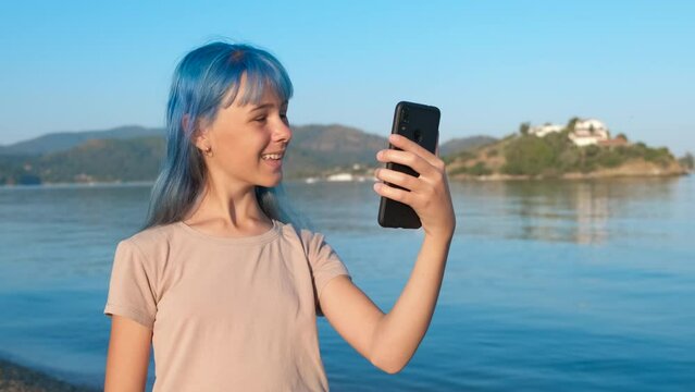 Blue teen speak on phone by sea. A blue haired young girl makes a video on the river bank during weekend in summer.