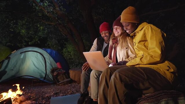 Three laughing people using computer during campsite sitting together on winter night by bonfire. Happy young group of joyful friends enjoying surfing net . Camping tent for outdoor nature getaway.