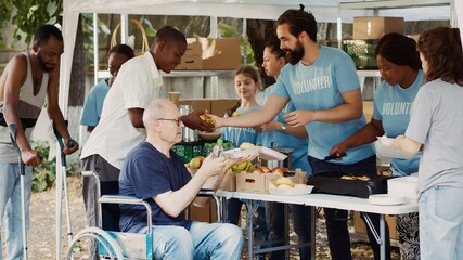 Charity organization shows devotion to fighting hunger and poverty by giving away free meals to homeless people. Warm food is provided by volunteers to caucasian wheelchair user and less privileged. - obrazy, fototapety, plakaty