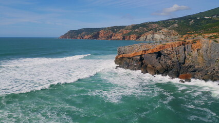 Sea hitting rugged coastal cliffs sunny day. Rough ocean waves rolling to rocks.