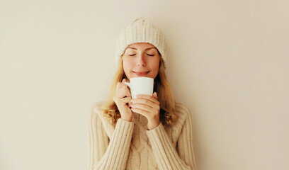 Winter portrait of happy satisfied young woman enjoying cup of hot tasty coffee or tea in warm knitted sweater and hat in early morning at home