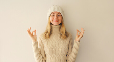 Portrait of relaxed caucasian young woman meditates in yoga position with folded arms and closed eyes wearing warm knitted sweater and hat on beige background