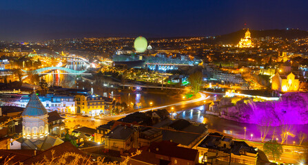 Fototapeta na wymiar Night spring cityscape of historical area of Tbilisi illuminated by colorful lights with view of modern bow-shaped bridge across Mtkvari River, Georgia