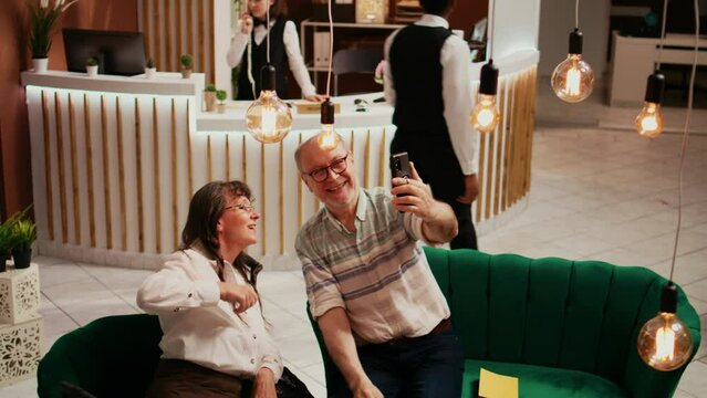 Elderly people taking photos in lobby, recording all activities on retirement vacation abroad. Old husband and wife take pictures on smartphone, making memories for family members.
