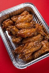 Grilled Chicken Wings in Spicy Teriyaki Sauce with Sesame Seeds in a Disposable Foil Box for Delivery on a Red Background