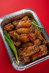 Grilled Chicken Wings in Spicy Teriyaki Sauce with Sesame Seeds in a Disposable Foil Box for Delivery on a Red Background