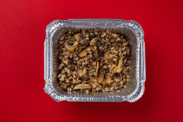 Buckwheat Porridge with Mushrooms, Champignons for Delivery in a Disposable Foil Box on a Red Background