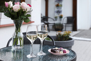 Vase with roses, glasses of wine and candies on glass table near house on outdoor terrace
