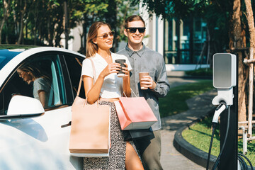 Young couple travel with EV electric car charging in green sustainable city outdoor garden in...