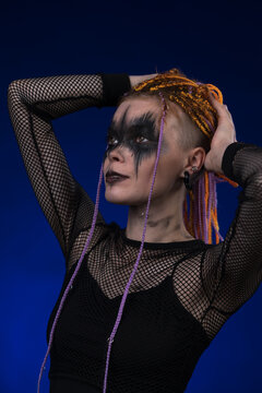 Cinematic portrait of young woman with orange color braids hairdo and horror black stage make-up painted on face. Studio shot on blue background. Part of photo series