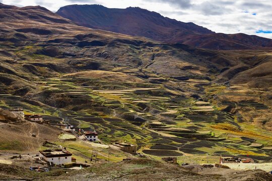 The landscape near Hikkim in Spiti Valley displays rust-colored mountains contrasted with green terraced fields. Traditional houses and prayer flags hint at the area's spiritual significance.