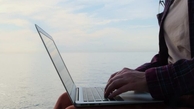 Static shot close-up woman's hands typing on a laptop keyboard by the sea. Female freelancer working on the seashore. Remote work, digital nomad concept.