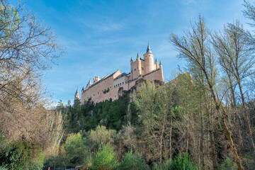Fototapeta na wymiar Castillo de cuento europeo en lo alto de una colina, rodeado de vegetación. El Alcázar de Segovia visto desde el mirador del valle, un día soleado con cielo azul.