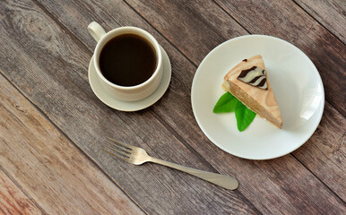 A plate with a slice of fresh creamy cake with mint leaves, a fork and a cup of hot coffee on a light wooden table.