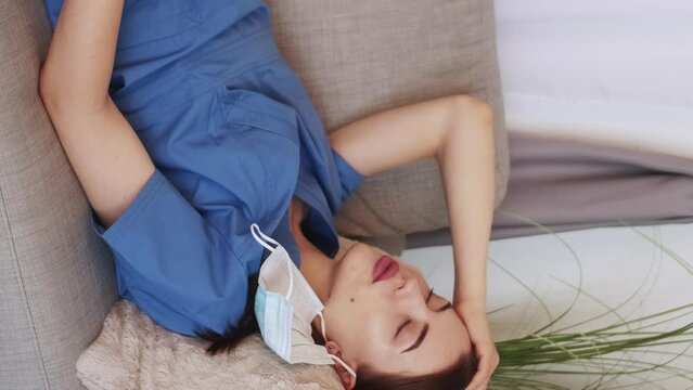 Vertical video. Exhausted doctor. Medical profession. Tired woman lying on couch in staff room taking off protective mask light room interior.