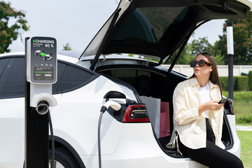 Young woman use smartphone to pay for electricity at public EV car charging station green city...