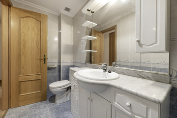 Bathroom of a home with two-tone white and blue tiles divided by a marine border, frameless mirror on the wall and white porcelain sink and marble countertop on white lacquered wooden furniture